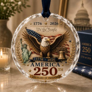 May include: A circular, faceted glass ornament featuring a bald eagle with wings spread, the Statue of Liberty, and the Liberty Bell. It displays the text 1776-2026, We The People, America 250th, and 250 Years of Freedom, suspended by a dark blue ribbon.