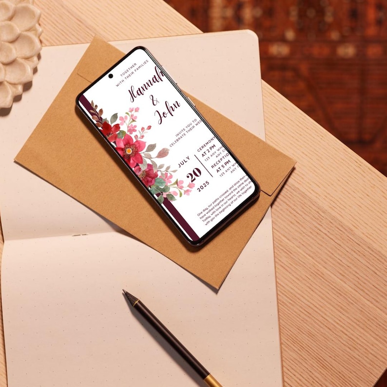 K&ouml;nnte beinhalten: Ein Smartphone zeigt eine Hochzeitseinladung mit den Namen "Hannah & John" und Veranstaltungsdetails. Die Einladung hat rote und rosa Blumendekorationen. Das Telefon liegt auf einem braunen Umschlag, auf einem Notizbuch, mit einem Stift daneben.