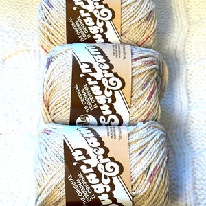 May include: Three skeins of Sugar'n Cream yarn in a speckled beige color, with flecks of pink, purple, and blue. Each skein has a brown label with the brand name and text. The yarn is arranged vertically on a white, textured surface.