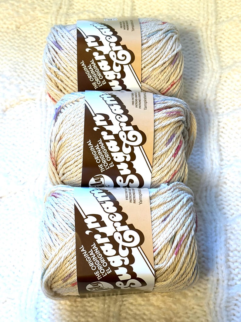 May include: Three skeins of Sugar'n Cream yarn in a speckled beige color, with flecks of pink, purple, and blue. Each skein has a brown label with the brand name and text. The yarn is arranged vertically on a white, textured surface.