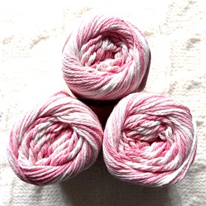 May include: Three balls of yarn in a soft pink and white striped pattern. The yarn is tightly wound, creating a spiral effect. The yarn is set against a textured white background, suggesting a craft or knitting project.