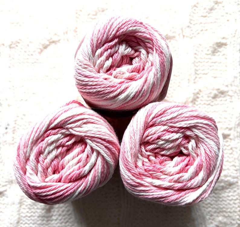 May include: Three balls of yarn in a soft pink and white striped pattern. The yarn is tightly wound, creating a spiral effect. The yarn is set against a textured white background, suggesting a craft or knitting project.