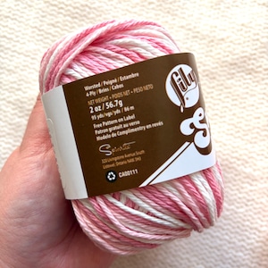 May include: A ball of Lily Sugar'n Cream yarn in shades of pink and white. The label indicates it is worsted weight, 2 oz (56.7g), and 95 yards (86 m). The label also includes the text "Free Pattern on Label".