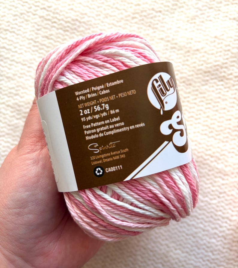 May include: A ball of Lily Sugar'n Cream yarn in shades of pink and white. The label indicates it is worsted weight, 2 oz (56.7g), and 95 yards (86 m). The label also includes the text "Free Pattern on Label".