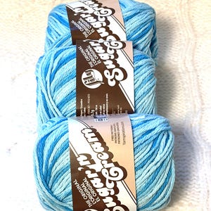 May include: Three skeins of blue and white variegated Sugar'n Cream yarn. Each skein has a label with the brand name and the words "The Original" in English, French, and Spanish. The yarn is displayed on a white knitted surface.