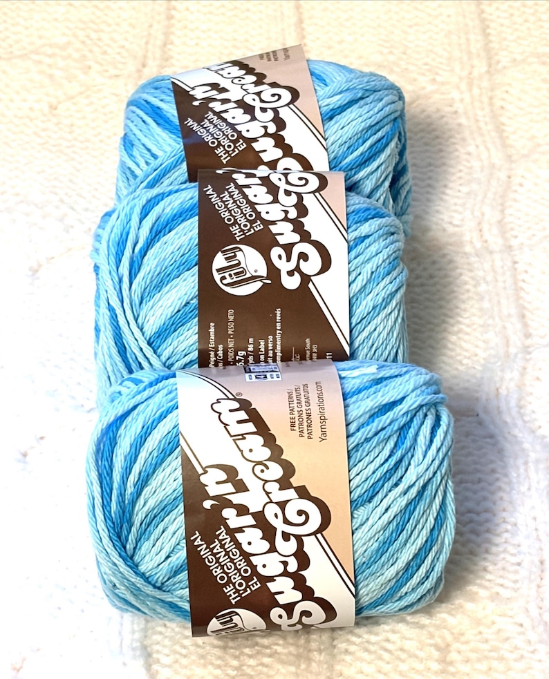 May include: Three skeins of blue and white variegated Sugar'n Cream yarn. Each skein has a label with the brand name and the words "The Original" in English, French, and Spanish. The yarn is displayed on a white knitted surface.