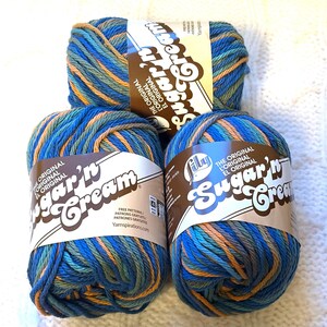 May include: Three skeins of Lily Sugar'n Cream yarn in shades of blue, green, and orange. Each skein has a brown label with the brand name and text that reads "THE ORIGINAL". The yarn is arranged on a white, textured surface.