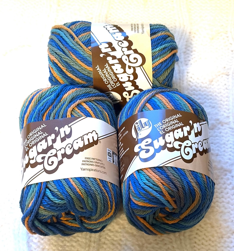 May include: Three skeins of Lily Sugar'n Cream yarn in shades of blue, green, and orange. Each skein has a brown label with the brand name and text that reads "THE ORIGINAL". The yarn is arranged on a white, textured surface.