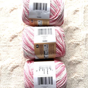 May include: Three skeins of yarn with a pink and white variegated pattern. Each skein has a label that reads "STRAWBERRY FRAISE FRESA 102002". The yarn is arranged vertically on a cream-colored, textured surface.