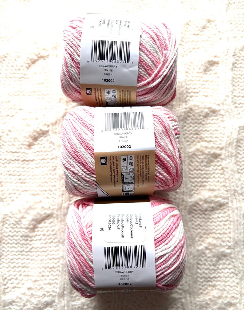 May include: Three skeins of yarn with a pink and white variegated pattern. Each skein has a label that reads "STRAWBERRY FRAISE FRESA 102002". The yarn is arranged vertically on a cream-colored, textured surface.