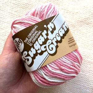 May include: A ball of pink and white Sugar'n Cream yarn. The yarn is wrapped with a brown label that reads "Sugar'n Cream" in a whimsical font. The label also includes the text "The Original" in English, French, and Spanish.