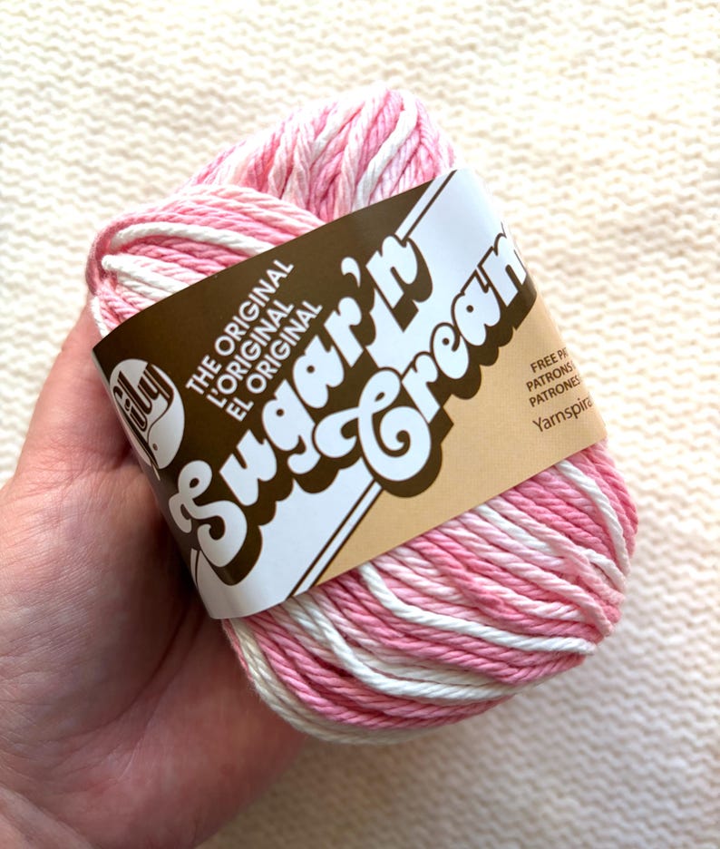 May include: A ball of pink and white Sugar'n Cream yarn. The yarn is wrapped with a brown label that reads "Sugar'n Cream" in a whimsical font. The label also includes the text "The Original" in English, French, and Spanish.