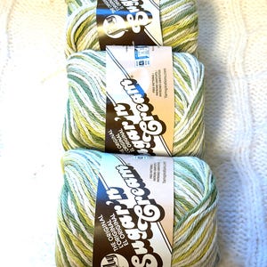 May include: Three skeins of Sugar'n Cream yarn in shades of green, yellow, and white. Each skein has a brown label with the brand name and the words "The Original" in English, French, and Spanish. The background is a white, textured surface.