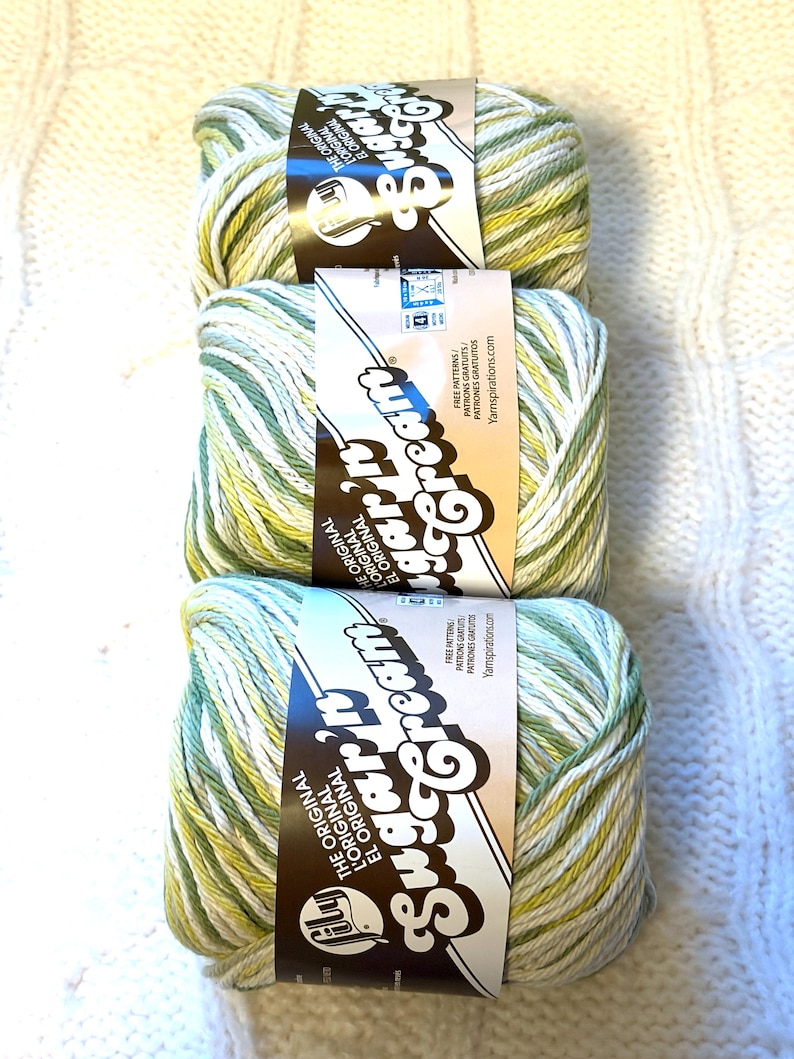 May include: Three skeins of Sugar'n Cream yarn in shades of green, yellow, and white. Each skein has a brown label with the brand name and the words "The Original" in English, French, and Spanish. The background is a white, textured surface.