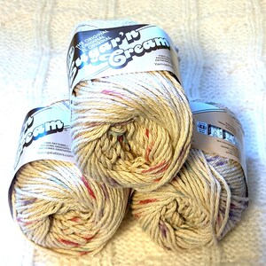 May include: Three skeins of Sugar'n Cream yarn in a speckled cream color with red, blue, and purple flecks. The yarn is labeled with the text "Sugar'n Cream" and "The Original". The yarn is sitting on a white, textured surface.