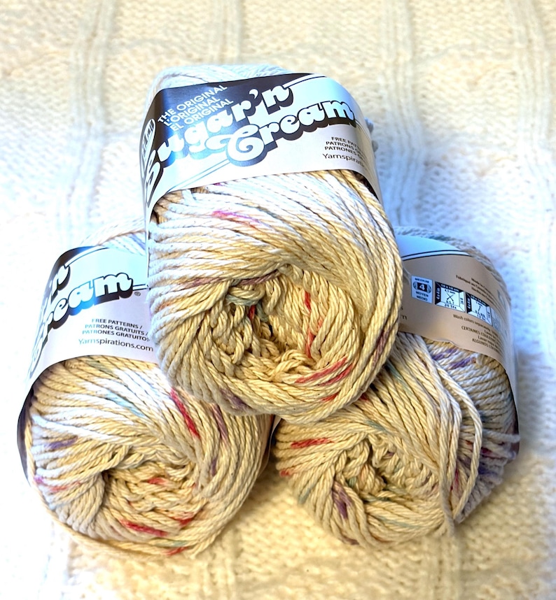 May include: Three skeins of Sugar'n Cream yarn in a speckled cream color with red, blue, and purple flecks. The yarn is labeled with the text "Sugar'n Cream" and "The Original". The yarn is sitting on a white, textured surface.