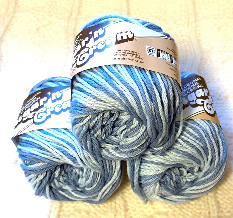 May include: Three skeins of Sugar'n Cream yarn in shades of blue and white. The yarn is labeled with the brand name and information about free patterns. The yarn is suitable for knitting and crochet projects.