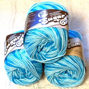 May include: Three skeins of blue and turquoise variegated yarn. Each skein has a label that reads "Sugar'n Cream" and "The Original" in English and Spanish. The yarn is arranged on a white, textured surface.