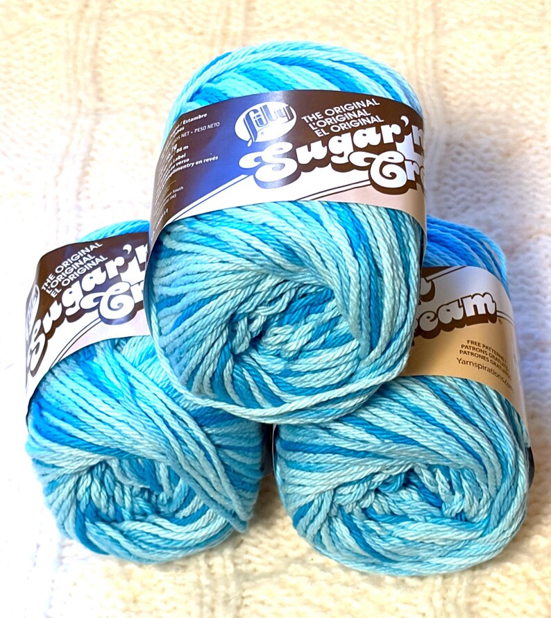 May include: Three skeins of blue and turquoise variegated yarn. Each skein has a label that reads "Sugar'n Cream" and "The Original" in English and Spanish. The yarn is arranged on a white, textured surface.