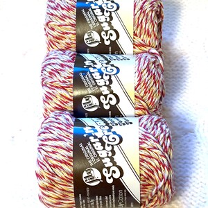May include: Three skeins of Lily Sugar'n Cream cotton yarn. The yarn is a mix of red, white, and yellow. Each skein has a black and white label with the brand name and the words "THE ORIGINAL" in white.