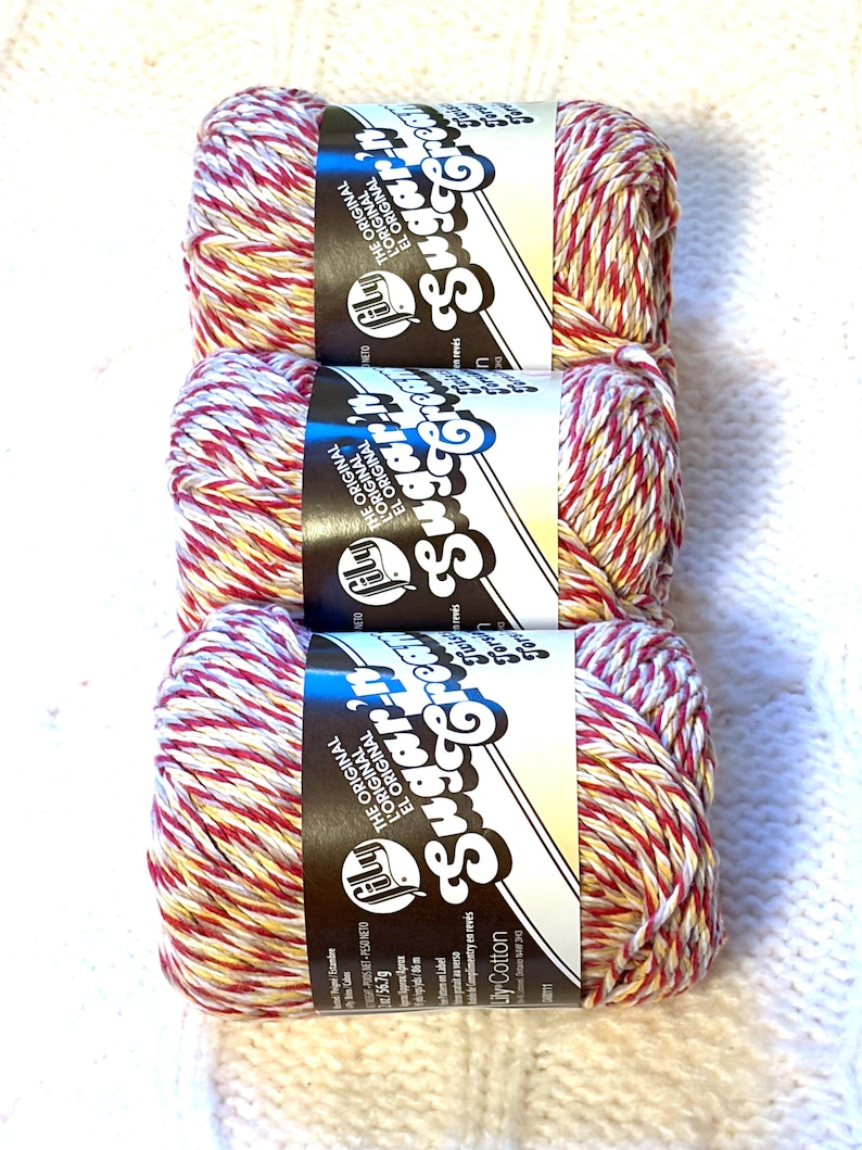May include: Three skeins of Lily Sugar'n Cream cotton yarn. The yarn is a mix of red, white, and yellow. Each skein has a black and white label with the brand name and the words "THE ORIGINAL" in white.