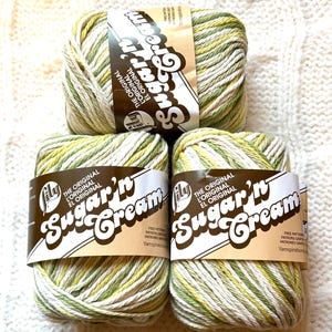 May include: Four skeins of Lily Sugar'n Cream yarn in a variegated green, yellow, and white colorway. The yarn is labeled with the brand name and product details. The skeins are stacked on a white knitted surface.