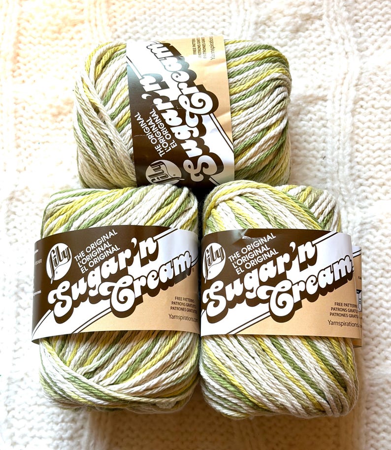 May include: Four skeins of Lily Sugar'n Cream yarn in a variegated green, yellow, and white colorway. The yarn is labeled with the brand name and product details. The skeins are stacked on a white knitted surface.