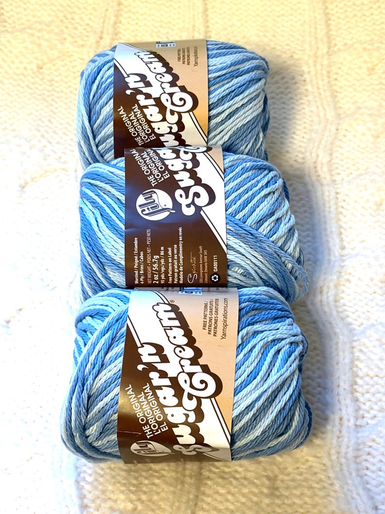 May include: Three skeins of blue variegated yarn, each with a brown label featuring the text "Sugar'n Cream". The yarn is a blend of light and dark blues, perfect for knitting or crocheting projects. The labels also include the text "The Original".