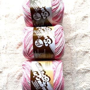 May include: Three skeins of pink and white striped yarn with brown labels. The labels read "Sugar Cream" and "The Original". The yarn is arranged vertically on a textured, off-white surface, ideal for knitting or crochet projects.