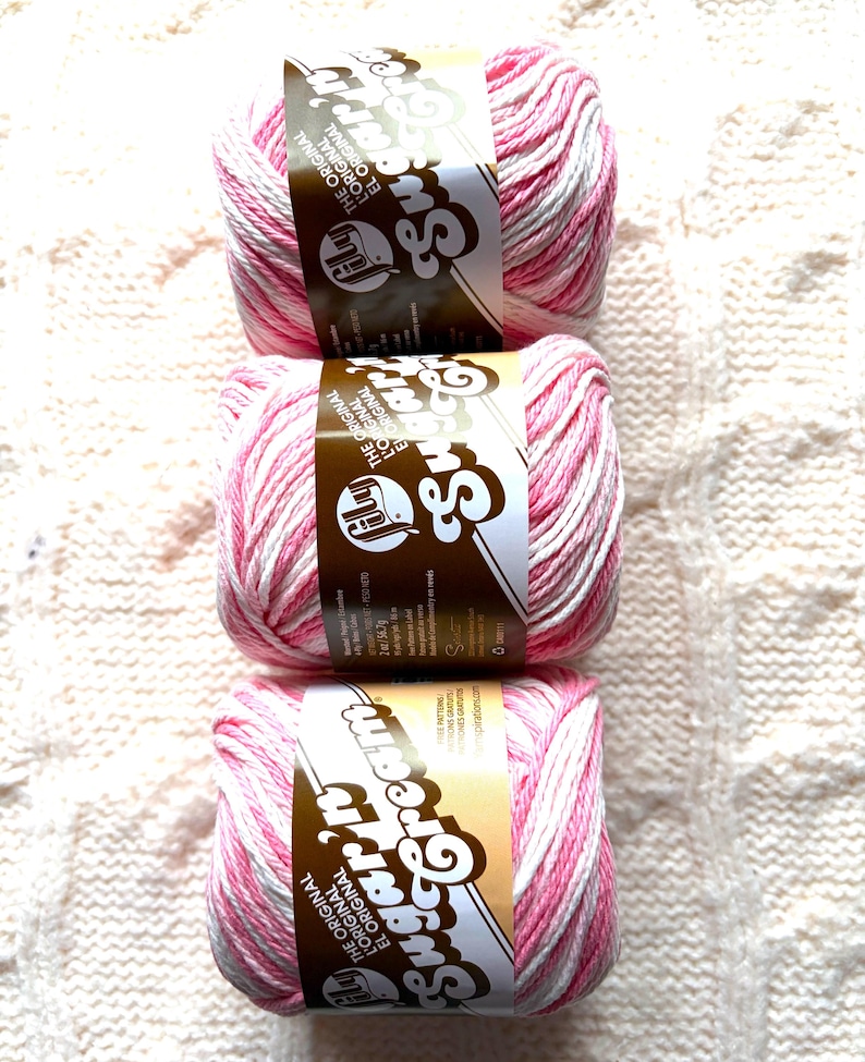 May include: Three skeins of pink and white striped yarn with brown labels. The labels read "Sugar Cream" and "The Original". The yarn is arranged vertically on a textured, off-white surface, ideal for knitting or crochet projects.