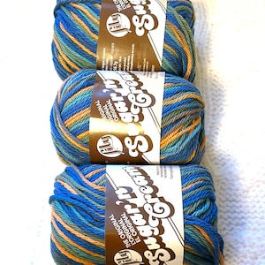 May include: Three skeins of Sugar'n Cream yarn in shades of blue, orange, and green. Each skein has a brown label with white text. The yarn is arranged vertically on a white, textured surface, ideal for knitting and crochet projects.