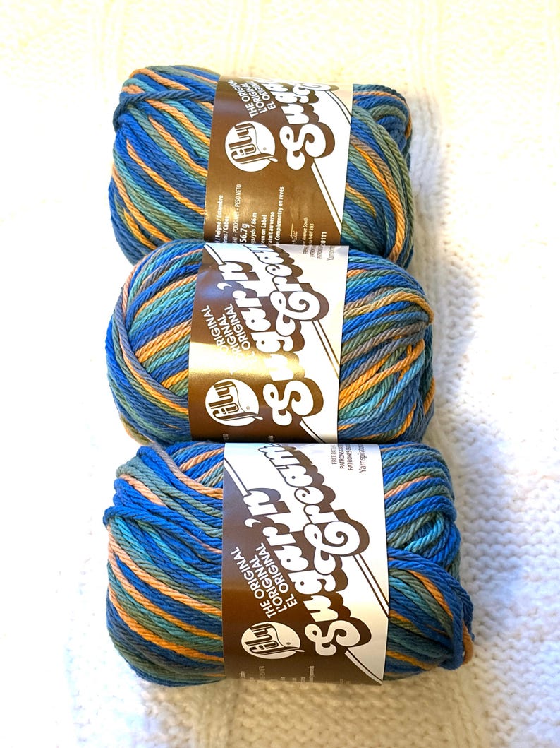 May include: Three skeins of Sugar'n Cream yarn in shades of blue, orange, and green. Each skein has a brown label with white text. The yarn is arranged vertically on a white, textured surface, ideal for knitting and crochet projects.