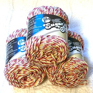 May include: Four spools of Lily Sugar'n Cream cotton yarn. The yarn is a mix of red, yellow, and white. The labels are blue and white with the brand name and product details. The yarn is stacked on a white, textured surface.