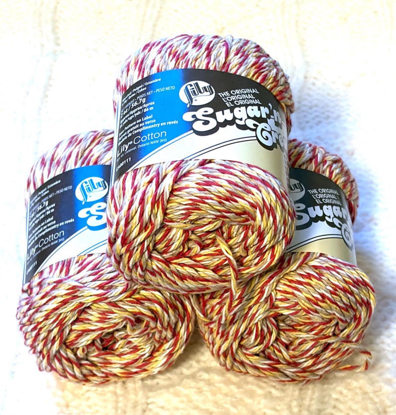 May include: Four spools of Lily Sugar'n Cream cotton yarn. The yarn is a mix of red, yellow, and white. The labels are blue and white with the brand name and product details. The yarn is stacked on a white, textured surface.