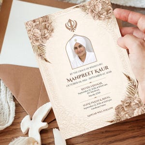 May include: A beige invitation card with floral and leaf accents, featuring a portrait and text. The text includes a name, dates, and event details. The card is held near a brown envelope.