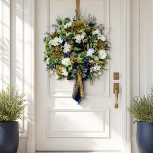 May include: A floral wreath with white, green, and blue flowers, and a plaid ribbon, hangs on a white door. The wreath is suspended by a gold-colored ribbon. A brass door handle and two potted plants flank the door.