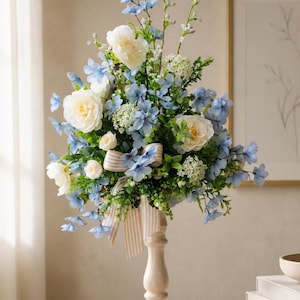 May include: A floral arrangement featuring white peonies, light blue blossoms, and green foliage set in a tall, light-colored wooden pedestal vase. A beige and white striped ribbon is tied around the base of the flowers, adding a decorative touch to the centerpiece.