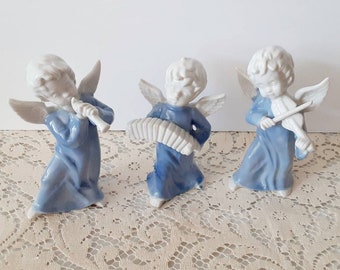 May include: Three porcelain angel figurines in a row, each playing a musical instrument. The angels wear blue robes and have white wings and hair. One plays a flute, one an accordion, and the third a violin.