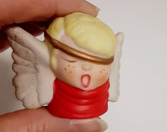 May include: A small porcelain angel figurine with white wings, a red robe, and a gold headband. The angel has blonde hair and a closed-eye expression.