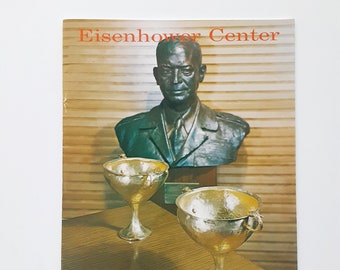 May include: A bronze bust of Dwight D. Eisenhower, former President of the United States, is displayed in front of a wood-paneled wall. Two gold chalices sit on a wooden table in the foreground. The text "Eisenhower Center" is printed in red at the top of the image.