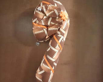 May include: A brown and white football-patterned pillow shaped like a candy cane. The pillow has orange ribbon accents and a small orange pom-pom on the top.