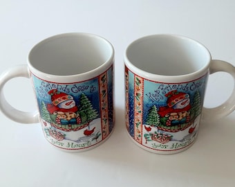 May include: Two white ceramic mugs featuring a colorful winter illustration of a snowman wearing a red hat and scarf, surrounded by pine trees and red birds. The text "Keep friends close to your heart" is printed in blue script on the front.