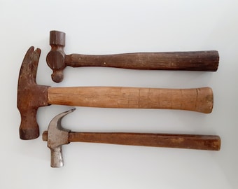 May include: Three vintage hammers with wooden handles. The hammers are in various states of wear and tear, with some rust and scratches. The hammers are arranged in a row on a white surface.