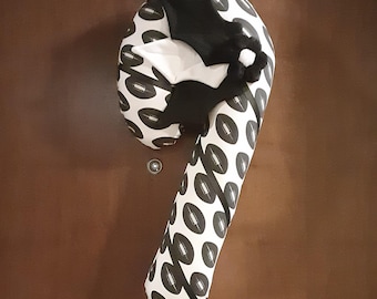 May include: A candy cane shaped door decoration featuring a black and white fabric pattern of American footballs. The top of the decoration is accented with a black fabric bat shape and a small black and white flower-like pom-pom embellishment.