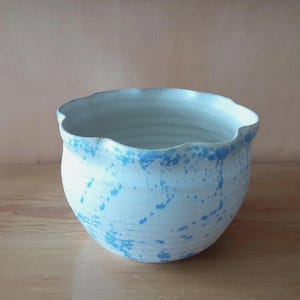 May include: A ceramic bowl with a light blue and white speckled design. The rim of the bowl has a wavy, irregular shape, and the interior is a solid light blue color. The bowl is sitting on a wooden surface.