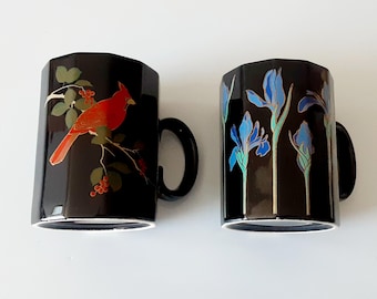 May include: Two black ceramic mugs with a faceted, octagonal shape. One mug features a red cardinal bird perched on a branch with green leaves and red berries. The other mug displays a pattern of blue iris flowers with green stems and leaves.