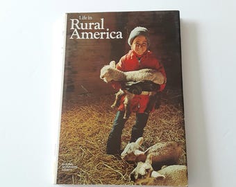 May include: A hardcover book titled Life in Rural America by the National Geographic Society. The cover features a photograph of a person wearing a red jacket and grey knit hat, holding a lamb in a barn with straw, with two other lambs nearby.