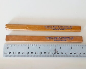 May include: Two flat, light brown wooden carpenter pencils with blue text reading MOORE LUMBER & HARDWARE, INC. BAILEY 838-5362 CASTLE ROCK 688-0397 ELIZABETH 546-4343. A silver metal ruler shows the pencils are approximately 7 inches long.