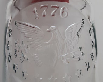 May include: A clear glass bottle featuring an embossed design of an eagle with outstretched wings, clutching arrows and an olive branch. The number 1776 is embossed above the eagle, surrounded by a circular arrangement of thirteen five-pointed stars.