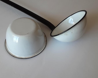 May include: A white enamel ladle with a long black handle and a matching white enamel bowl positioned upside down. Both pieces feature a thin black rim along the edges, showcasing a classic two-tone design with a smooth, glossy finish.